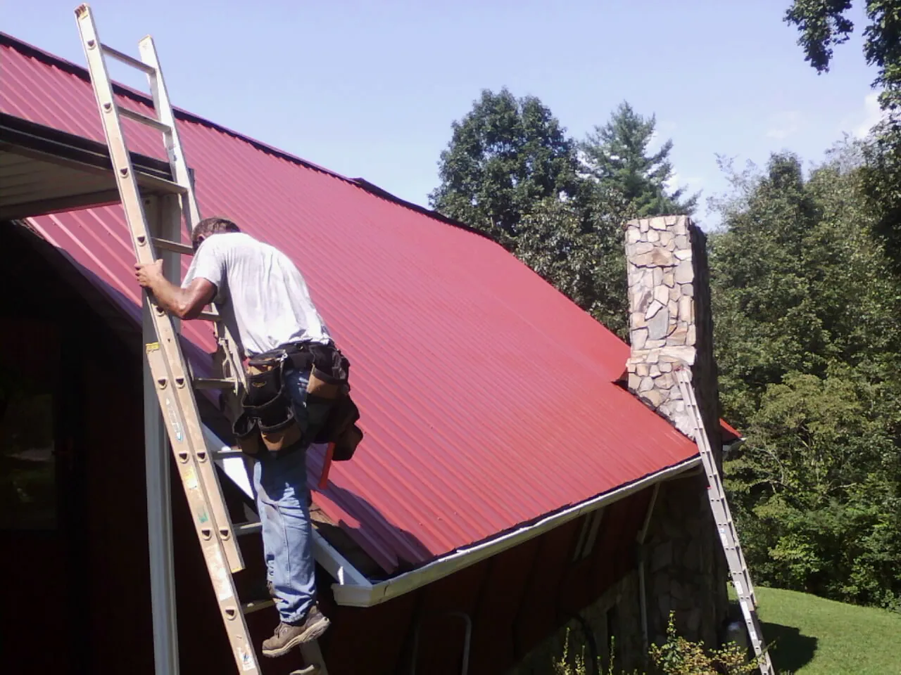 Roofer on a ladder beside a red metal roof and stone chimney — Soffit & Fascia Repair by Roofing Brothers in Hoyt